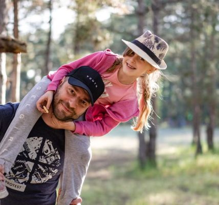 A joyful father giving his daughter a piggyback ride in a sunny forest setting.