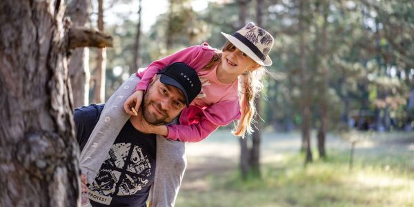 A joyful father giving his daughter a piggyback ride in a sunny forest setting.